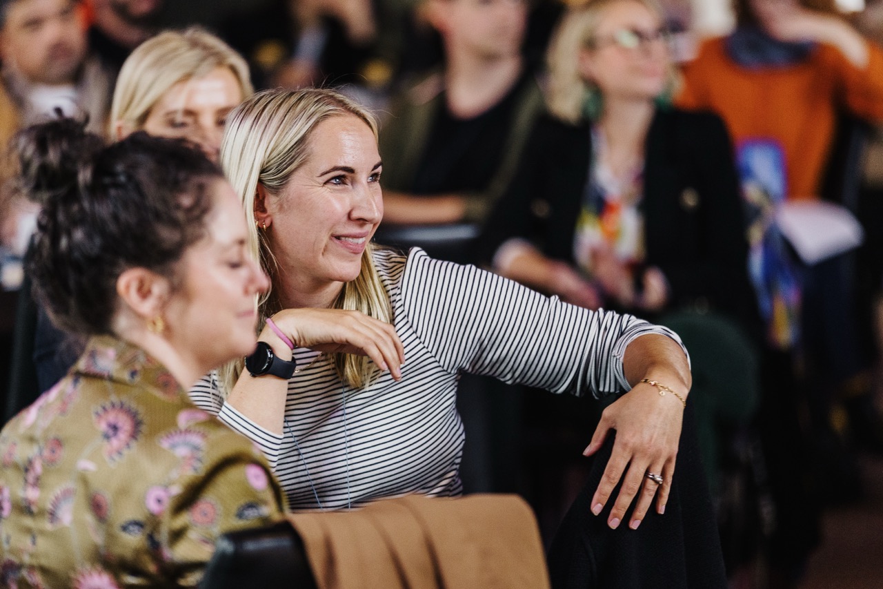 A woman with blonde hair wearing a striped shirt, smiling and sitting in a crowd at Design Leadership