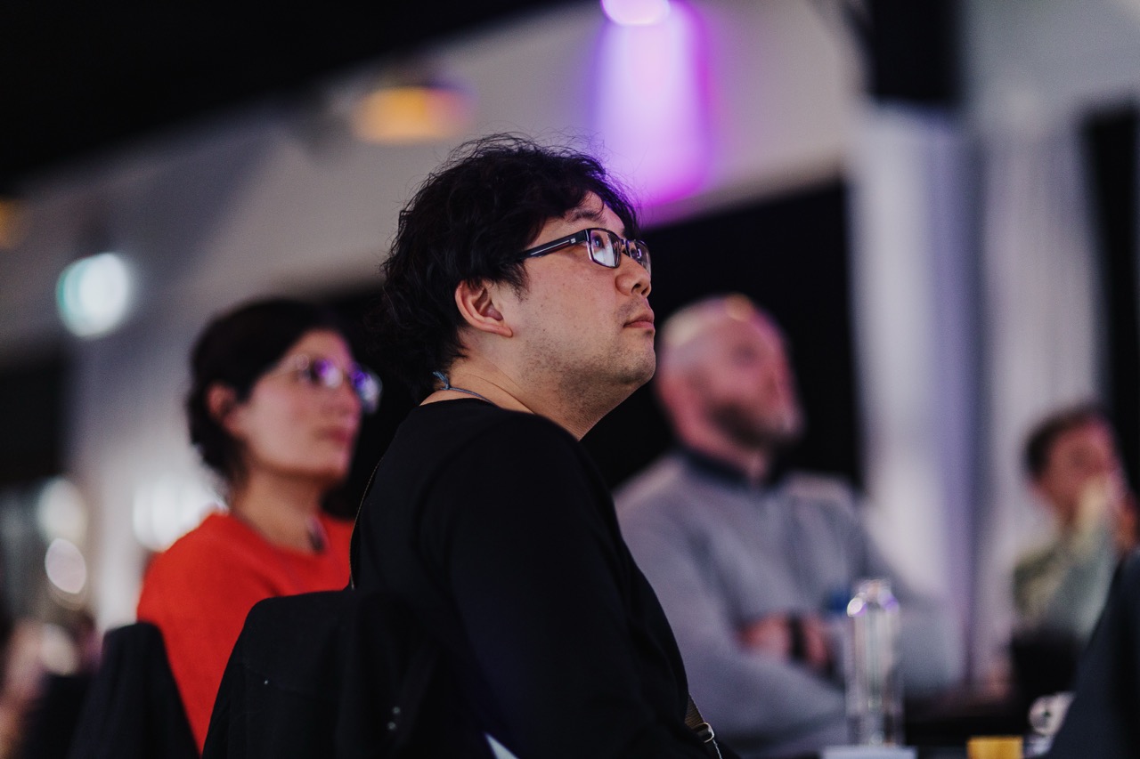 A man wearing glasses and a black shirt attentively listening during the Design Leadership event, with other attendees visible in the background