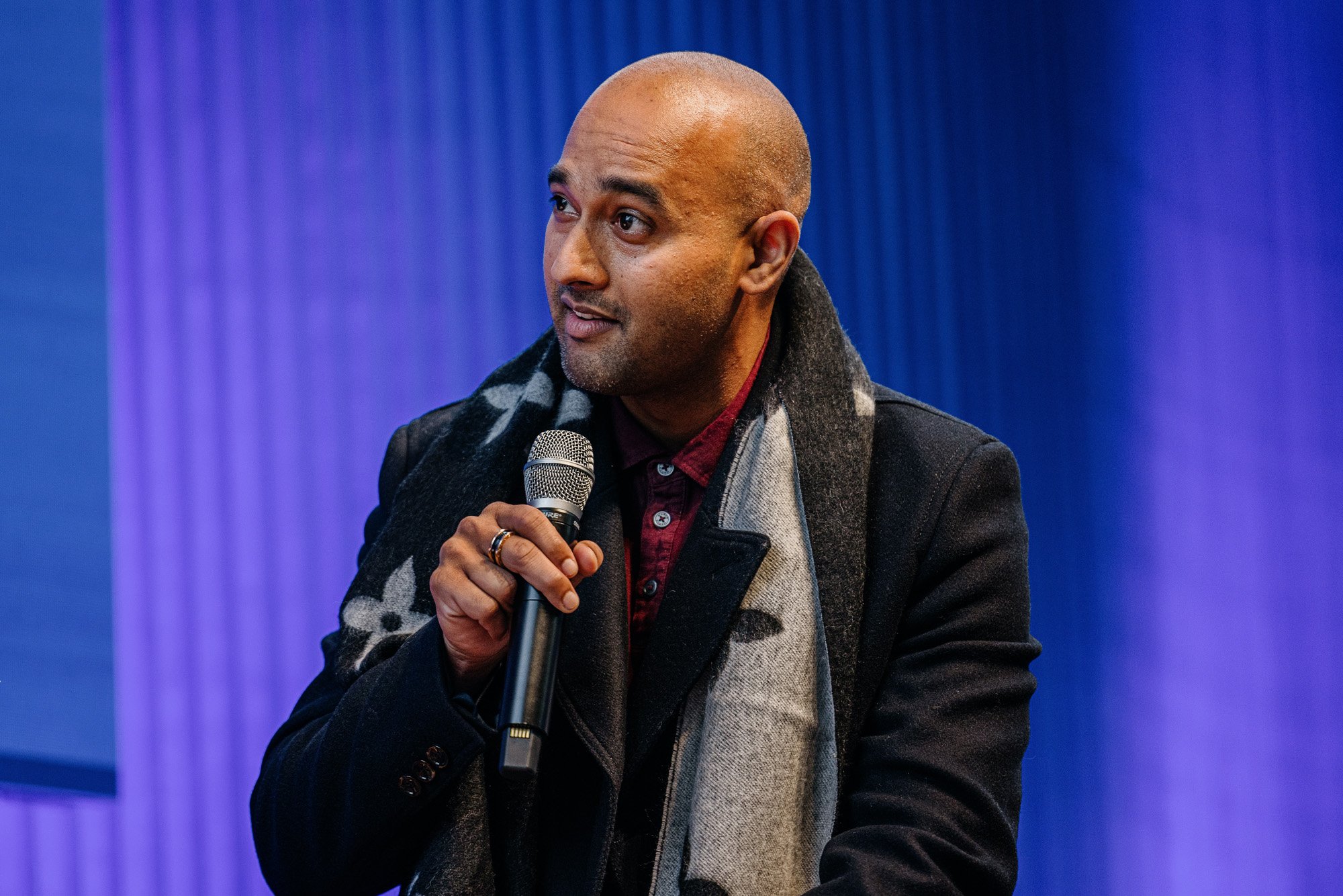 Abi Ayer peaking into a microphone. He is wearing a dark coat and a patterned scarf, with a purple backdrop behind him