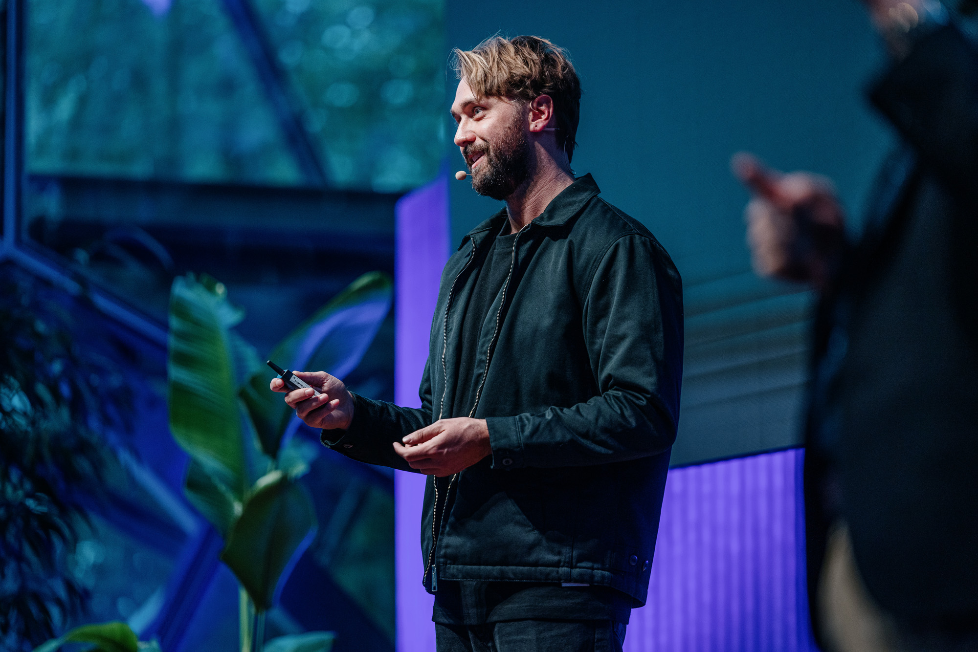 Oliver Ree giving a presentation at DO22, wearing a black jacket and using a microphone headset, with a leafy plant and large windows in the background