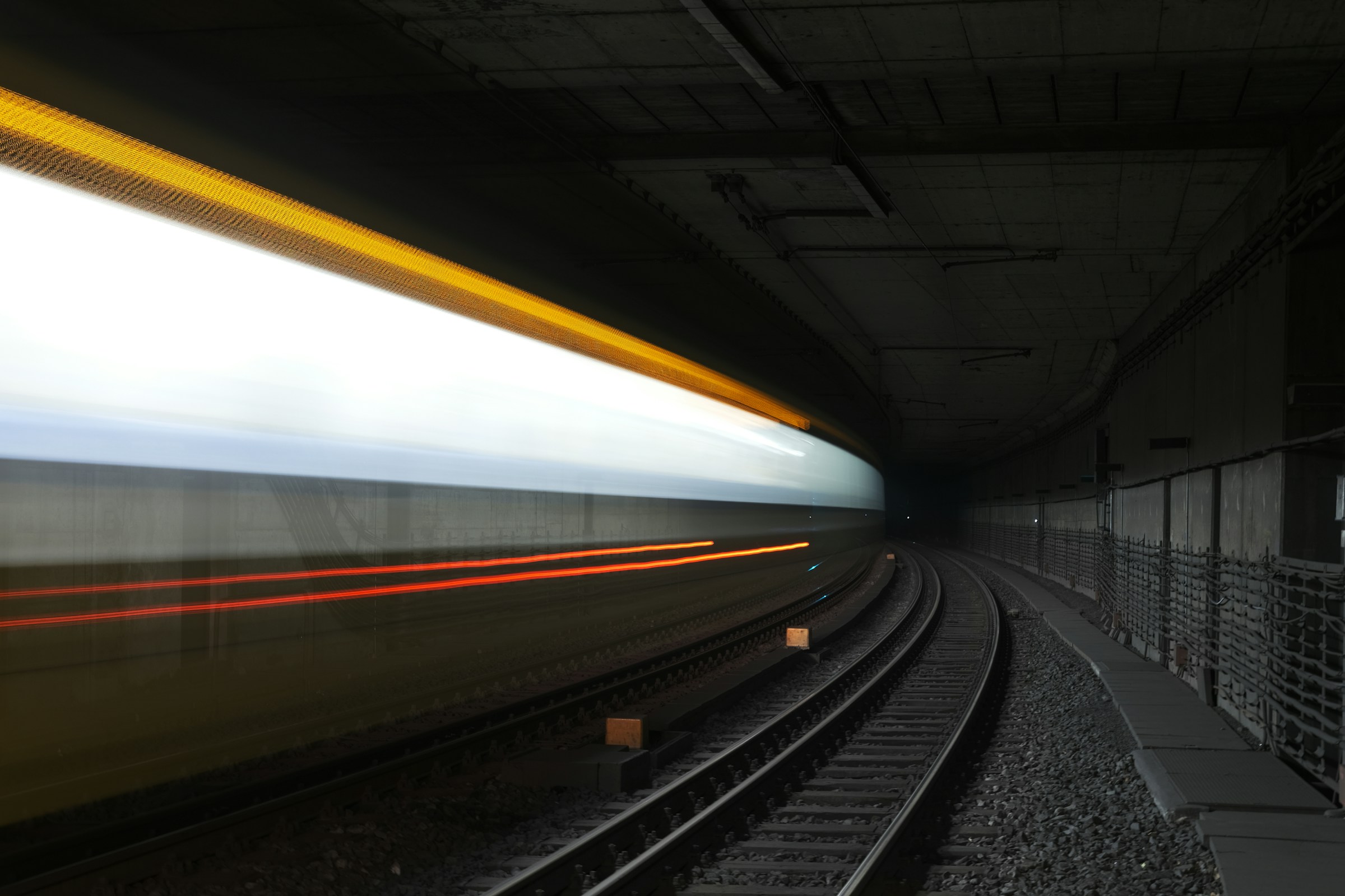 A long-exposure shot of a train speeding through a dark underground tunnel, creating blurred light streaks in white, yellow, and red. The train tracks curve into the distance, while the tunnel walls and ceiling are visible in the background.