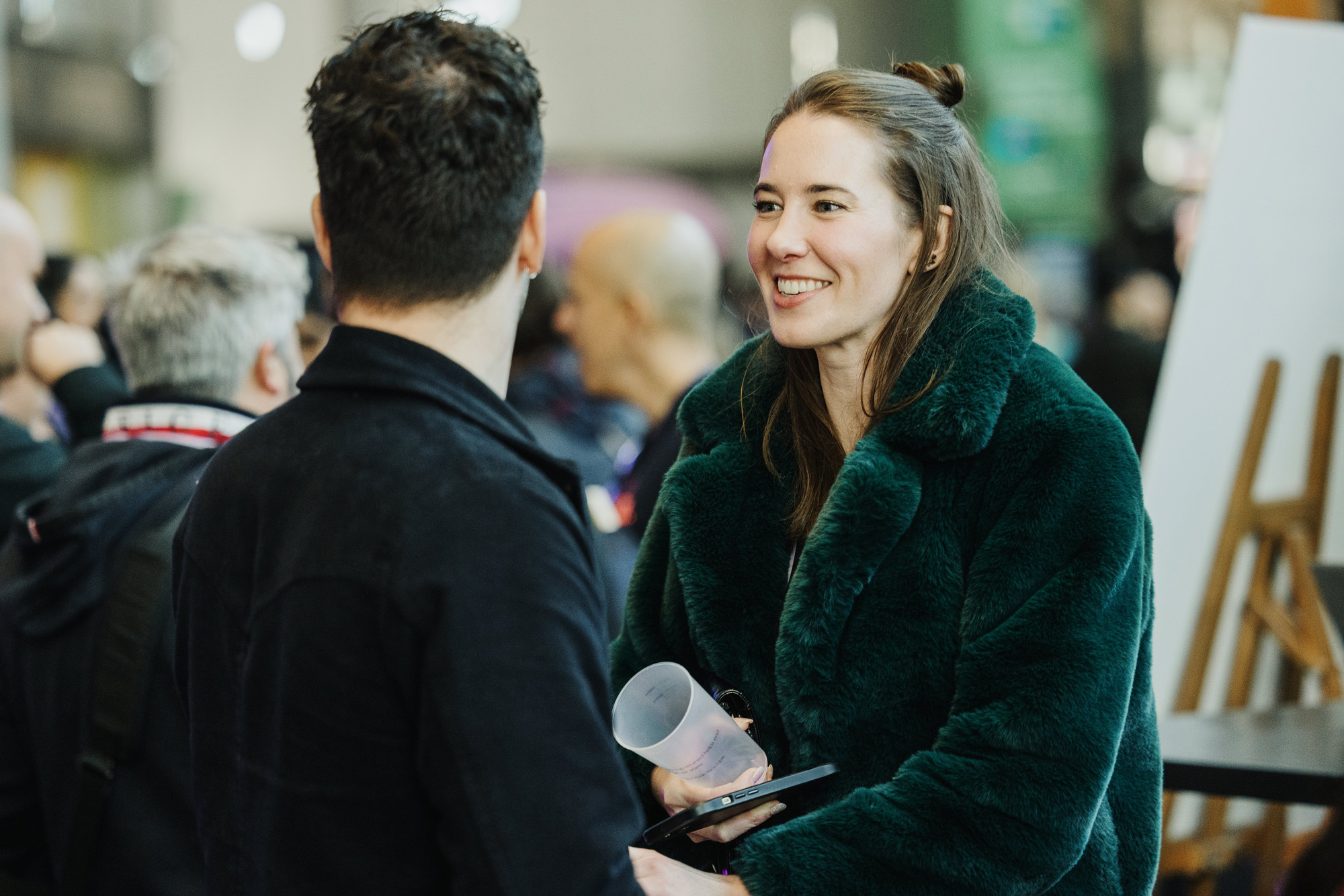 A candid moment at a Design Outlook event, showing a woman in a green faux fur coat smiling and engaging in conversation with a man dressed in a dark coat. The setting appears lively, with blurred attendees in the background creating a bustling atmosphere.