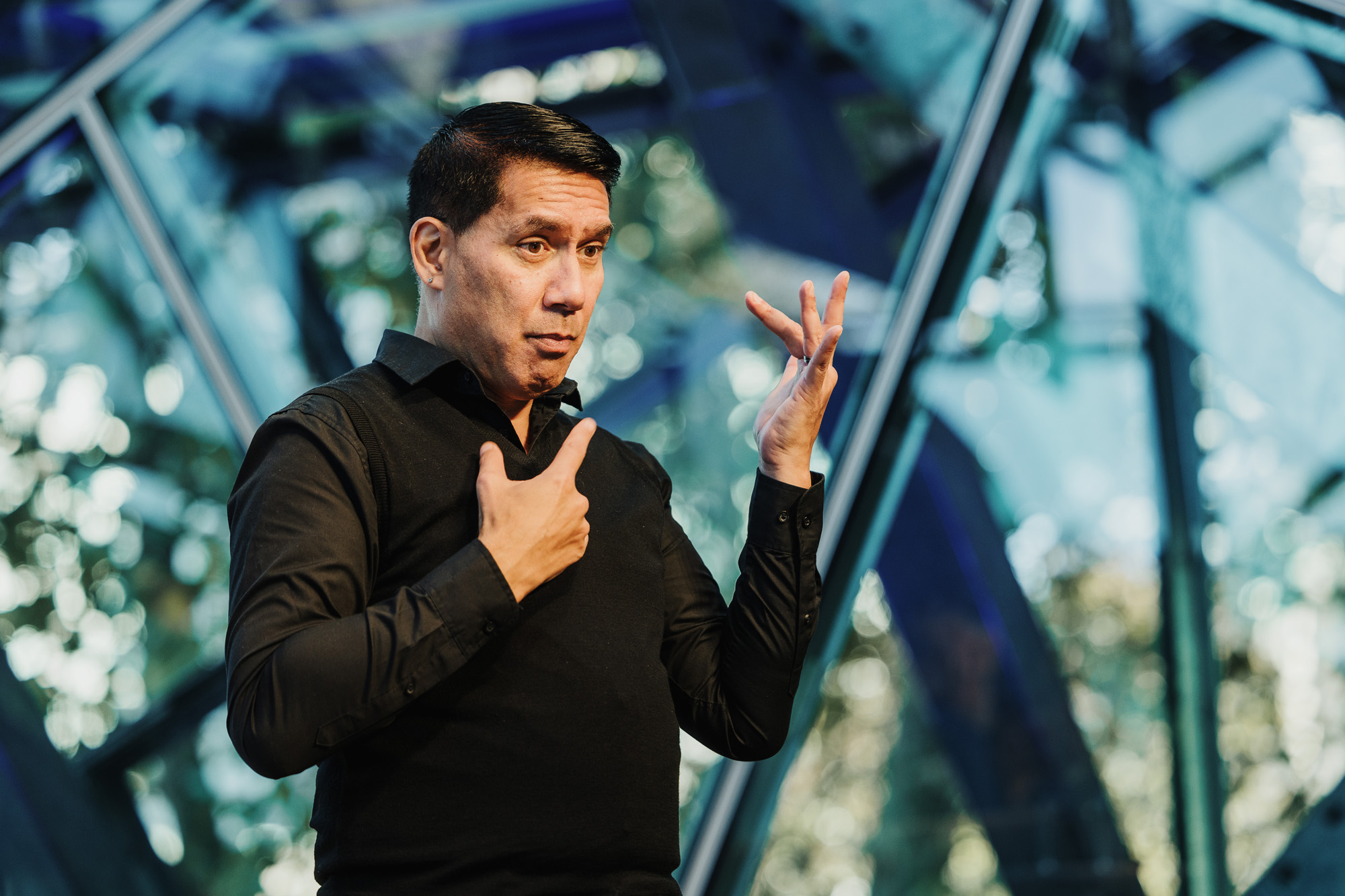 Marc, an Auslan interpreter, actively signing during a Design Outlook event. He is wearing a black shirt and vest, with expressive gestures and a focused expression. The background features modern glass architecture with natural light filtering through, adding to the professional atmosphere of the event