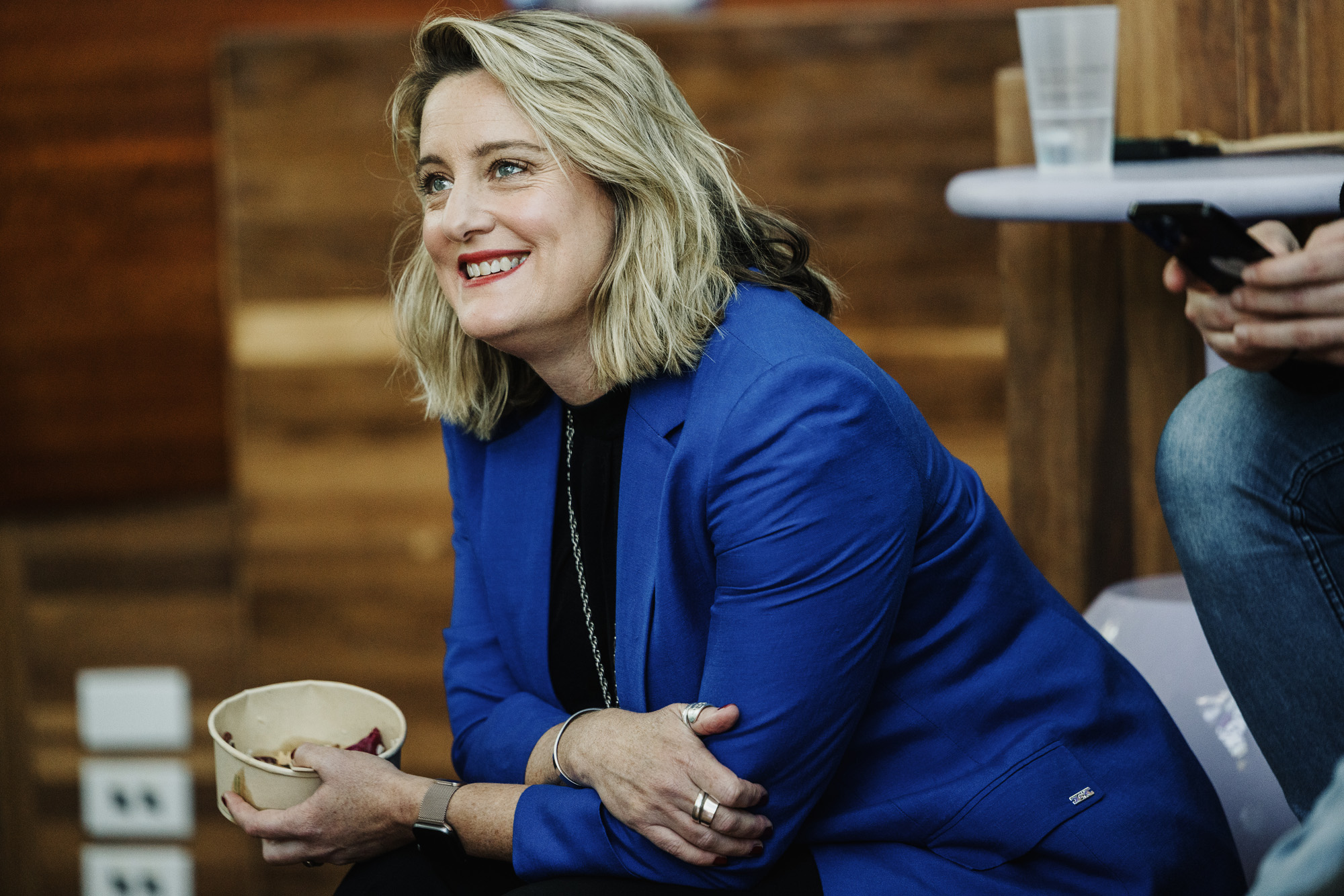 Trudi Boatwright wearing a vibrant blue blazer, seated and smiling warmly during a Design Outlook event. She is holding a bowl in her hands, appearing relaxed and engaged in the surroundings. The background features wooden textures and casual seating, enhancing the informal and welcoming atmosphere