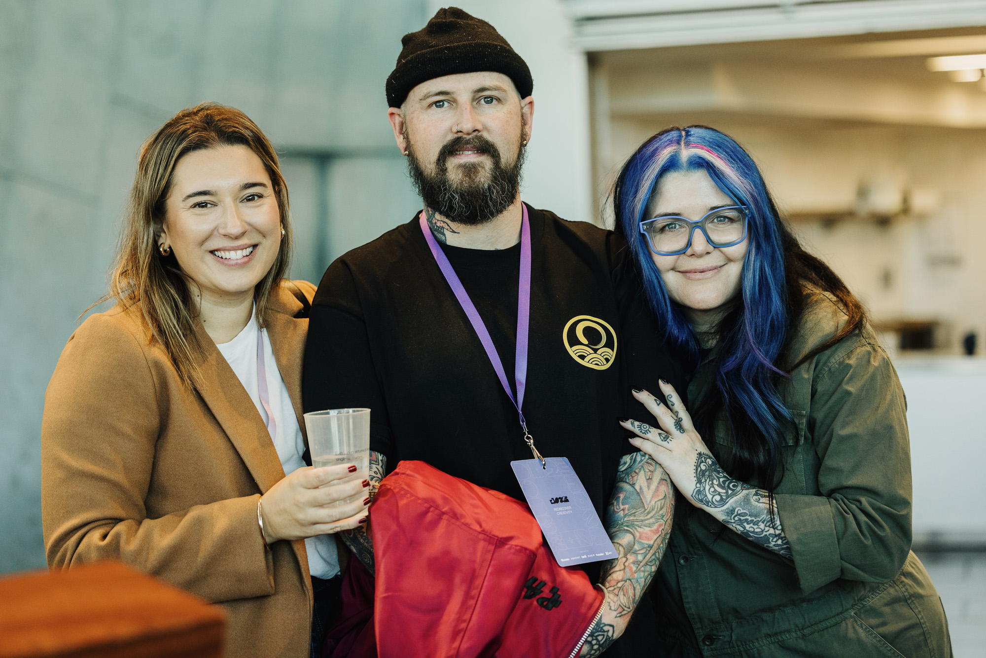 Three individuals posing together at a Design Outlook event, radiating camaraderie. The person on the left is smiling brightly, wearing a tan coat and holding a clear cup. In the center, a bearded individual wearing a black beanie, a black shirt with a gold emblem, and a purple lanyard holds a red jacket. On the right, Chi Ryan, with blue and black hair, blue glasses, and tattoos, leans in with a warm smile. The setting has a casual indoor atmosphere with soft lighting.