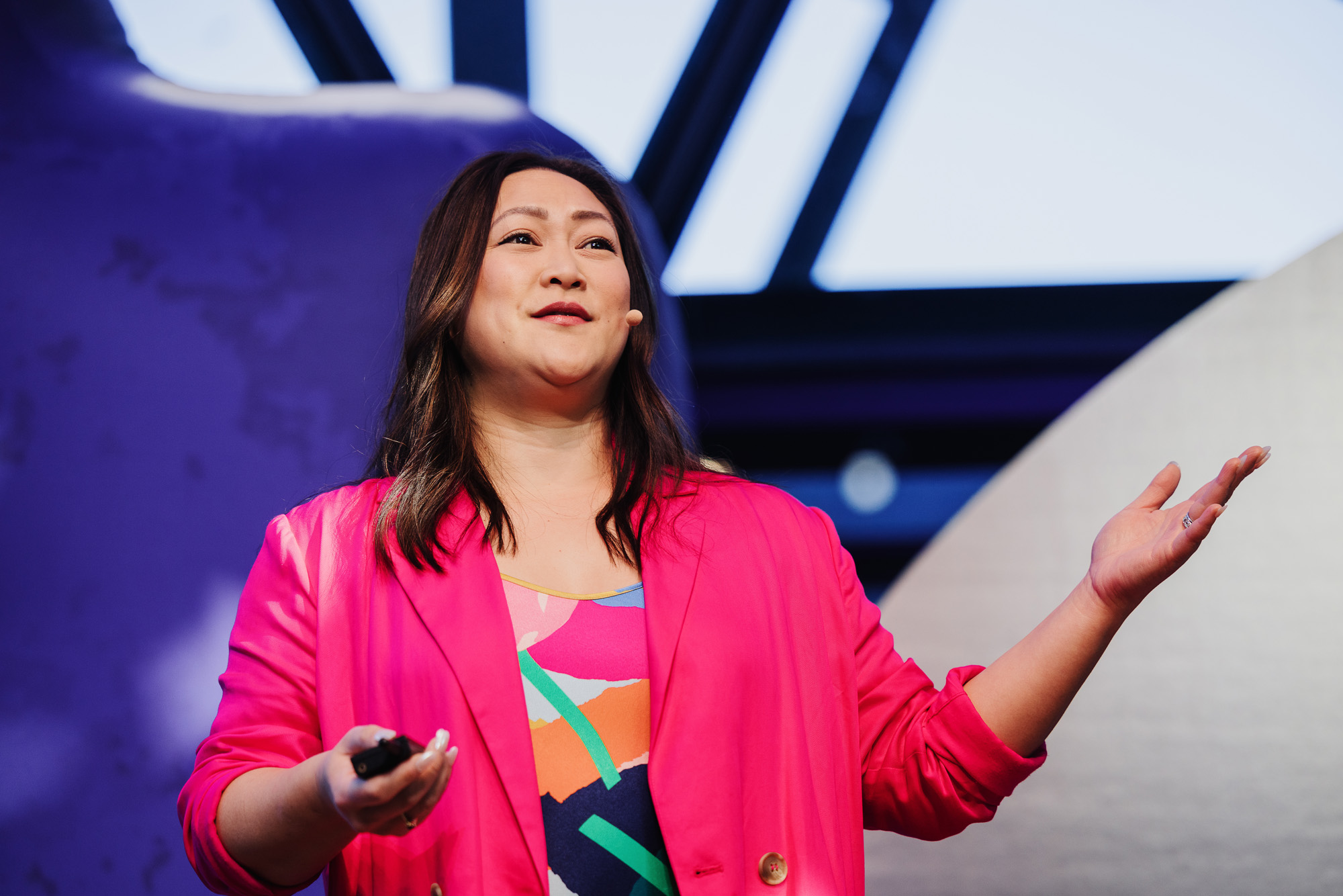 Jeanette Cheah presenting at a Design Outlook event, exuding confidence and enthusiasm. She is wearing a vibrant pink blazer over a colorful patterned top, holding a clicker in one hand while gesturing with the other. The background features abstract shapes and a modern architectural design, enhancing the lively and engaging atmosphere of the event.