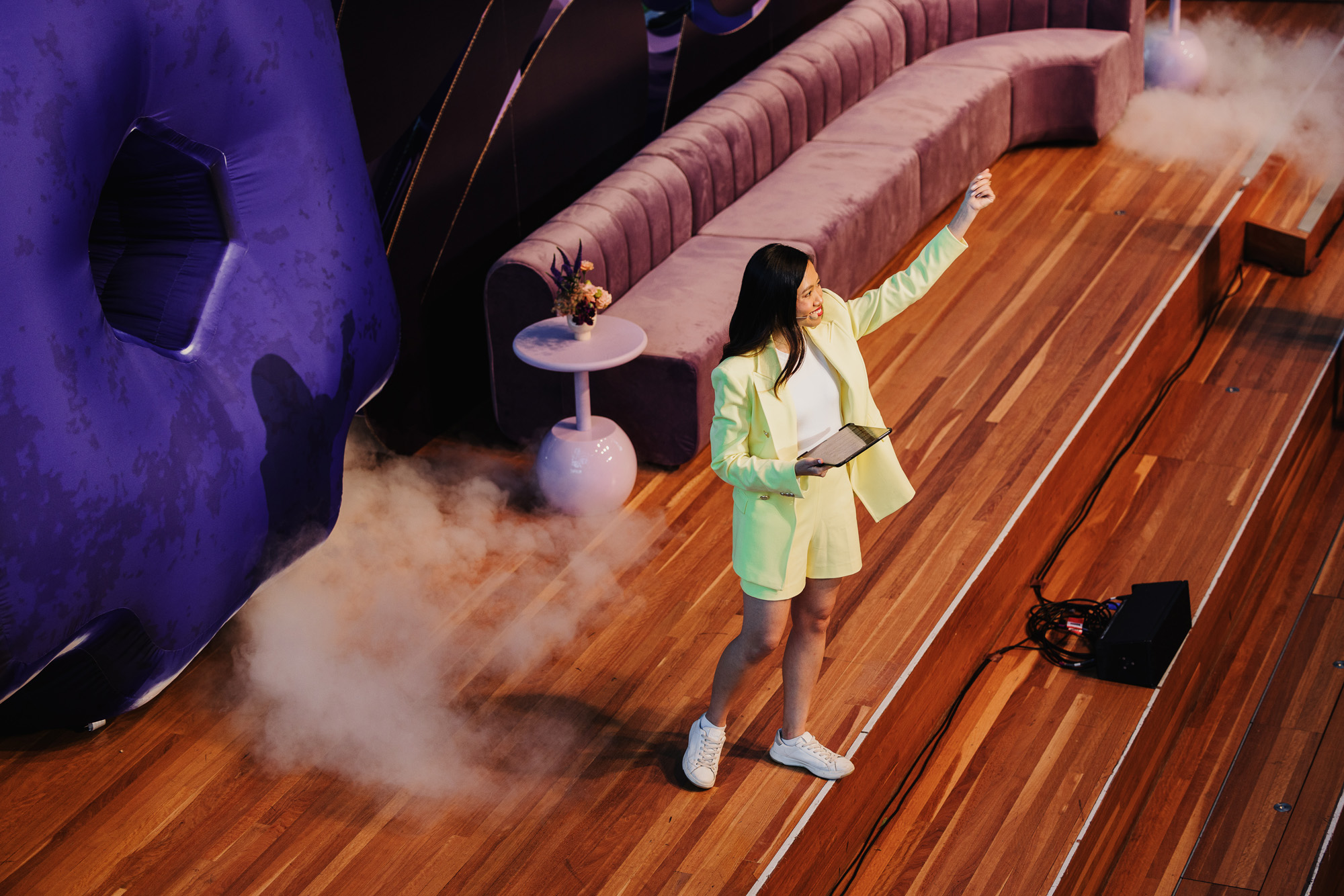 Berlin Liew presenting at a Design Outlook event, captured from an elevated angle. She is wearing a stylish light yellow blazer and shorts with white sneakers, holding a tablet in one hand while gesturing confidently with the other. The stage features a purple sofa, decorative side tables, and atmospheric smoke effects, with wooden floors and a modern, dynamic setup enhancing the scene.