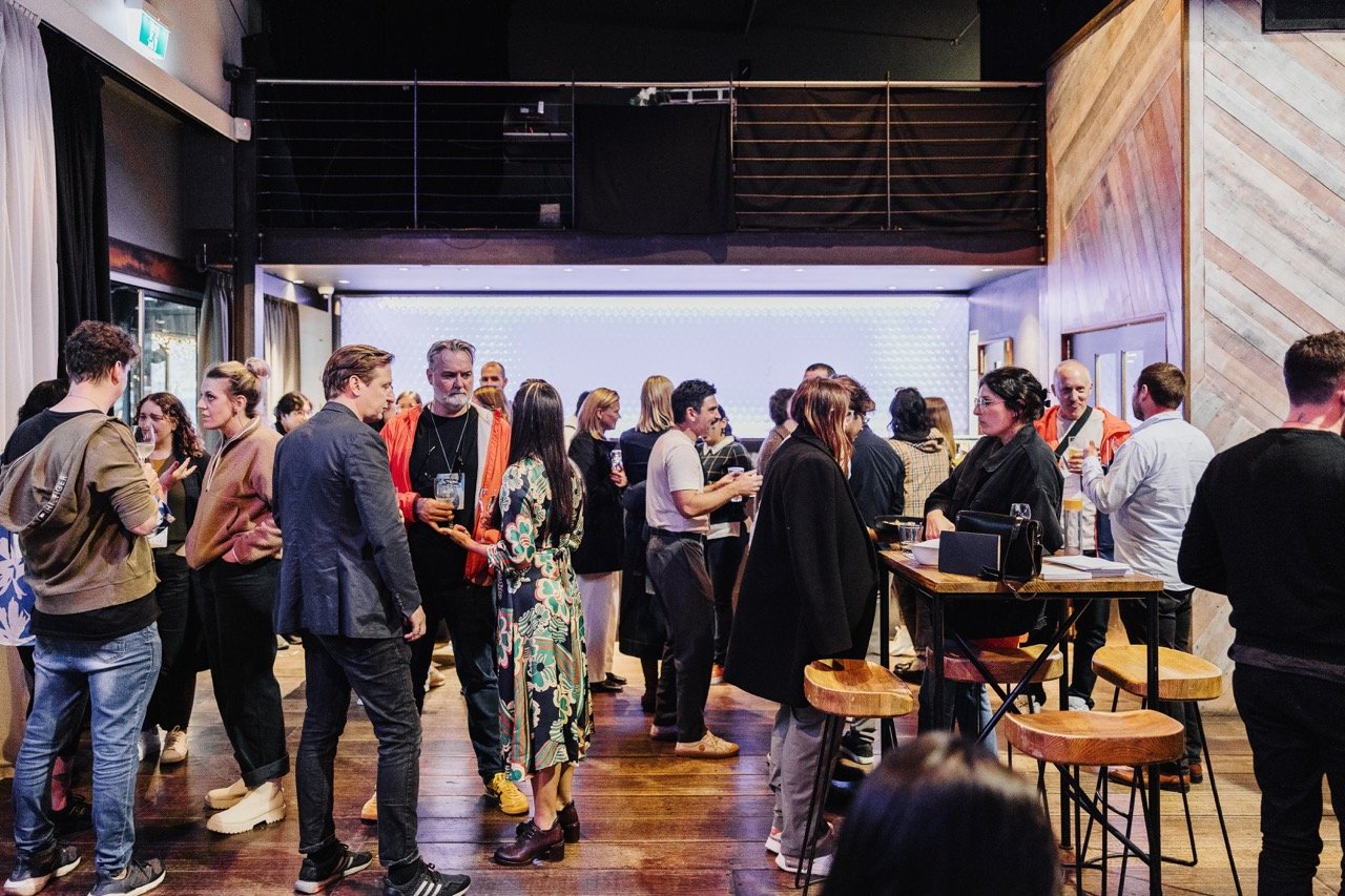 Guests networking during drinks after an event, engaging in lively conversations in a stylish indoor venue. The space features warm wooden accents, high-top tables with stools, and a softly lit backdrop, creating a relaxed and collaborative atmosphere to conclude the event.