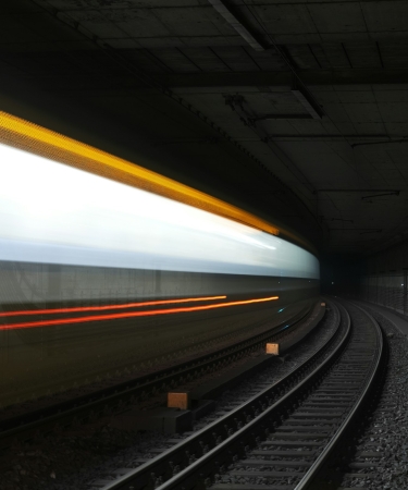 A long-exposure shot of a train speeding through a dark underground tunnel, creating blurred light streaks in white, yellow, and red. The train tracks curve into the distance, while the tunnel walls and ceiling are visible in the background.