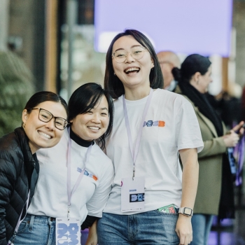 Three volunteers at the “DO23” conference smile as they pose together. Dressed in matching white event t-shirts with lanyards and name tags, they stand in a lively area with other attendees and event decor in the background, reflecting the energetic and welcoming spirit of the conference