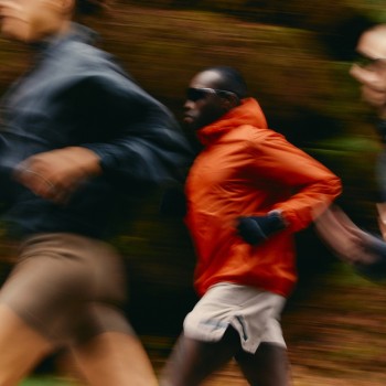 A dynamic, motion-blurred image of three runners moving through a forest trail. One runner in the center wears a bright orange jacket, standing out against the earthy tones of the background. The blurred motion emphasizes speed, energy, and the natural setting of the run.