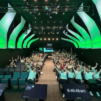 A full auditorium at UX Camp Melbourne, with attendees seated in rows under a ceiling featuring modern, angular green lighting. The audience, a mix of excited individuals wearing event t-shirts and casual clothing, raises their hands and smiles enthusiastically towards the camera, creating an energetic and welcoming atmosphere. In the foreground, two screens display the event logo and the current time, 9:46:48 am.