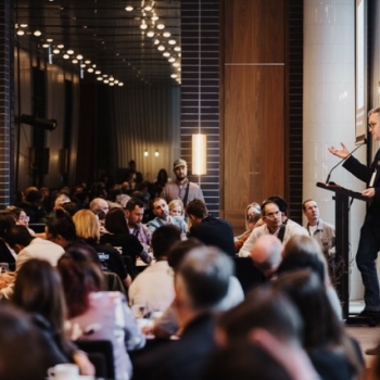 A speaker addresses a full room at a professional event. Attendees sit at round tables, listening closely in a modern, well-lit conference space.
