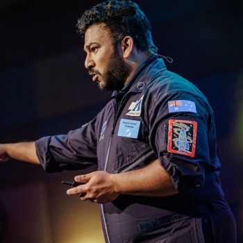 Dwayne Fernandes, wearing a space-themed flight suit, presenting passionately on stage at Accessibility & Inclusion 2025, gesturing with intensity.
