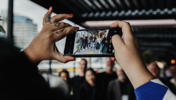 A close-up of a person’s hands holding a smartphone horizontally, capturing a group photo of people posing together in an outdoor covered area. The person’s hands feature bright red nail polish and tattoos of a sun and crescent moon on their fingers. The smartphone screen shows a clear image of the group, framed by blurred background elements in the real scene.