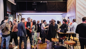 Guests networking during drinks after an event, engaging in lively conversations in a stylish indoor venue. The space features warm wooden accents, high-top tables with stools, and a softly lit backdrop, creating a relaxed and collaborative atmosphere to conclude the event.