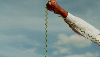 A hand holding a green vintage telephone receiver with a curly cord, extended against a backdrop of a blue sky with soft clouds. The receiver dangles from the hand, creating a whimsical, surreal effect in the open outdoor setting.