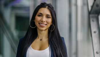 Emy Alegre smiling warmly, standing in front of a modern glass and metal architectural background. She is wearing a white top, a navy blazer, and large hoop earrings, projecting a professional and approachable demeanor.