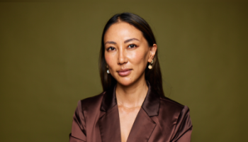 Rachel Zhang posing against a muted green background, wearing a satin brown blazer and pearl drop earrings. She has a calm and confident expression, exuding a professional and poised demeanor.