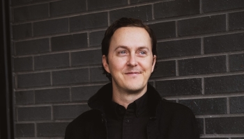 Ryan Rumsey standing against a dark gray brick wall, wearing a black shirt and jacket. He is looking slightly to the side with a relaxed smile, creating a professional yet approachable demeanor.