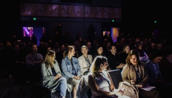 DO24 audience immersed in a session, with front-row attendees illuminated by stage lighting, engaged in conversation and note-taking in a dimly lit room.