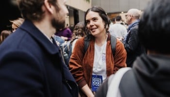 A DO24 attendee wearing a lanyard and backpack engages in a relaxed conversation amidst a bustling networking space filled with fellow participants.
