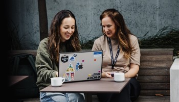 Two attendees collaborate outdoors at Design Leadership 2025, smiling over a laptop covered in stickers, with coffee cups resting on the table.