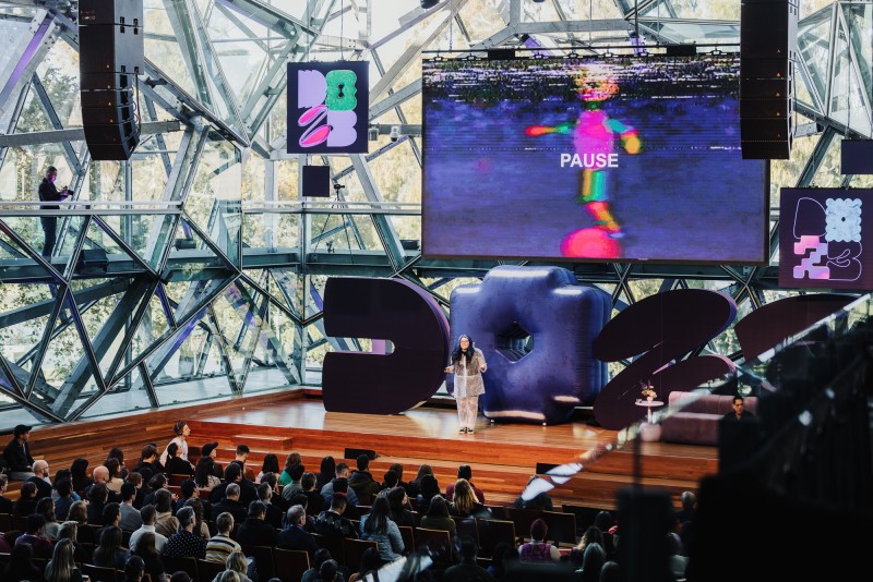 A photograph of Chi Ryan delivering the DO23 keynote presentation on a bight and vibrant stage
