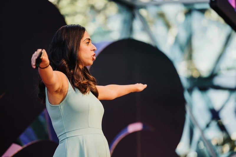 inita Israni presenting passionately at a Design Outlook event, captured mid-gesture with her arms extended. She is wearing a light teal dress, with the background showcasing abstract shapes and geometric architecture, adding a modern and dynamic atmosphere to the scene.