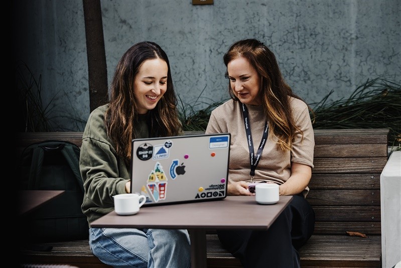 Two attendees collaborate outdoors at Design Leadership 2025, smiling over a laptop covered in stickers, with coffee cups resting on the table.