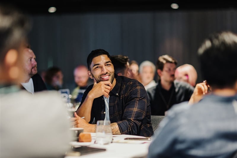 Aron Tucker smiling and listening during a roundtable discussion, seated at a table with water glasses and notebooks, surrounded by other attendees.