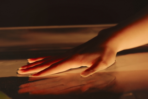 A close-up of a hand gently pressing against a smooth, reflective surface. Warm lighting casts a soft glow on the hand, creating subtle shadows and reflections.