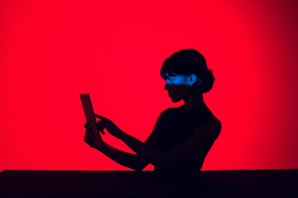 A silhouette of a person holding a tablet against a vibrant red background. The figure’s face and hands are partially illuminated by blue light, creating a striking contrast