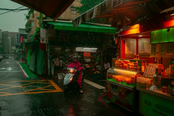 A rainy street scene in an urban market area, with a person wearing a pink raincoat riding a scooter. Brightly lit food stalls with colorful signs are on the right side, while green awnings cover parts of the shops in the background. Rain droplets are visible, adding to the atmosphere.