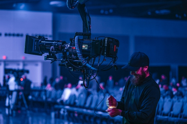 A camera operator in a dark room, wearing a black cap and working with a large professional camera rig suspended on a mechanical arm. The scene is lit with blue lighting, and rows of chairs with some blurred figures are visible in the background.