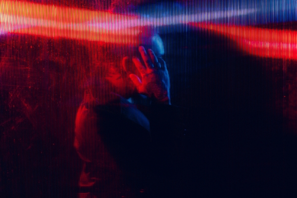 A mysterious figure seen through a translucent, ribbed surface with a hand raised, obscuring the face. The image is bathed in vibrant red and blue light, creating a dramatic, abstract effect.