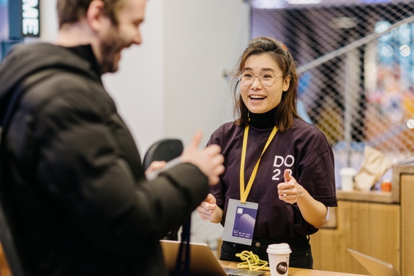 A volunteer at the “DO 2022” conference, wearing a purple shirt with the event logo, warmly engages with an attendee, gesturing enthusiastically. She is smiling, and there is a coffee cup on the table beside her. The attendee, a man in a dark jacket, listens attentively.