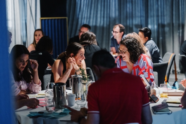 Attendees collaborating during a leadership event, seated around a table covered with materials like notebooks, sticky notes, and glasses of water. Two women in the foreground are smiling and engaged in animated conversation, creating a vibrant and interactive atmosphere. The background features other groups in discussion against softly lit curtains.