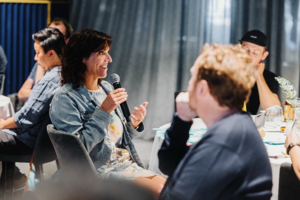 A woman seated at a table speaking into a microphone during a leadership event. She is smiling and wearing a denim jacket over a colorful outfit. Others at the table listen attentively, while the softly lit room and curtains in the background create a focused yet relaxed atmosphere.