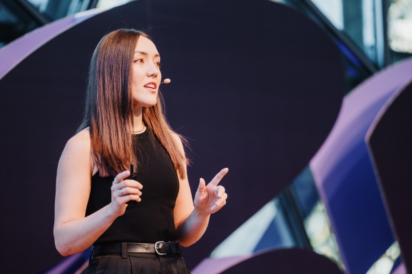 Jess Faccin presenting at a Design Outlook event, speaking with confidence while gesturing with her hands. She is dressed in a sleek black sleeveless top and black belt, with a professional and poised demeanor. The abstract purple and black stage design forms the backdrop, adding a modern touch to the setting.