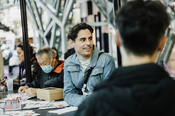 A young man wearing a denim jacket is seated at a table during a Design Outlook event, smiling and engaging in conversation with another person in the foreground. The table is covered with colorful materials and design elements, while other attendees are seen in the background, including an elderly man wearing a mask. The setting features modern architecture and a lively atmosphere