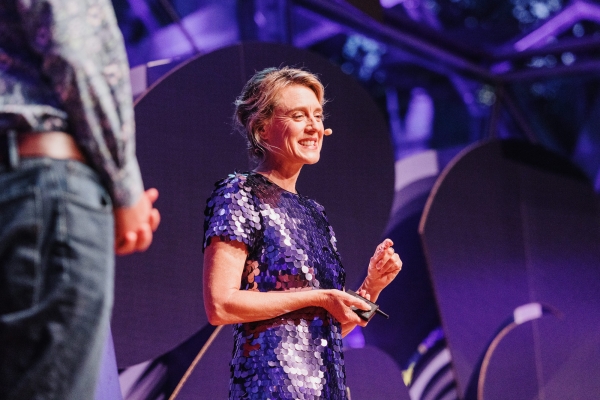 Dr. Barbara Doran presenting at a Design Outlook event, smiling confidently while speaking into a headset microphone. She is wearing a shimmering purple sequined dress, holding a clicker in one hand. The background features modern stage design elements illuminated with purple lighting, creating a vibrant and engaging atmosphere.