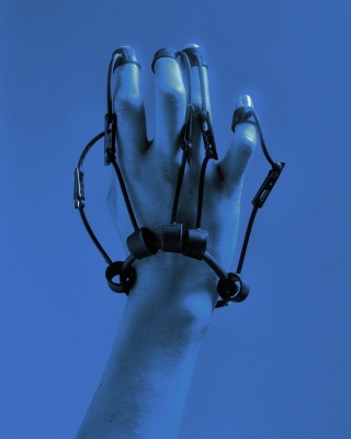 A hand wearing a complex structure of black straps and loops, attached to each finger, suggesting a mechanical or wearable tech device. The hand is illuminated in a cool blue tone against a matching blue background, creating a futuristic and experimental aesthetic. The straps and clips evoke themes of technology, augmentation, or control.