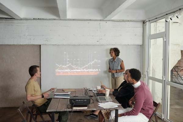 A group of colleagues in a casual meeting space, with a presenter standing next to a projected graph on the wall. Three team members are seated at a wooden table, listening attentively, with laptops and papers in front of them. The minimalist room features white walls, exposed beams, and large windows, creating a collaborative and modern work environment.