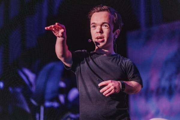 Joel, MC at DO24, on stage wearing a headset mic, gesturing with one hand while holding a remote, illuminated by dynamic purple-blue lighting.
