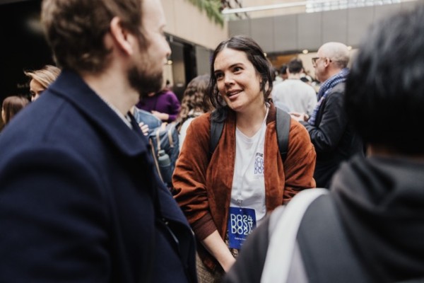 A DO24 attendee wearing a lanyard and backpack engages in a relaxed conversation amidst a bustling networking space filled with fellow participants.