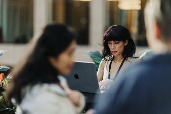 A DO24 attendee working intently on a laptop in an outdoor setting, framed by blurred figures in the foreground, capturing a moment of deep focus.