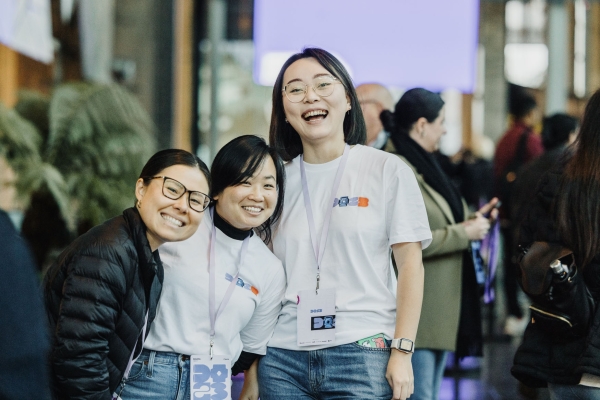 Three smiling Design Outlook 2023 volunteers wearing event T-shirts and lanyards, posing together in a busy venue space with attendees behind them.