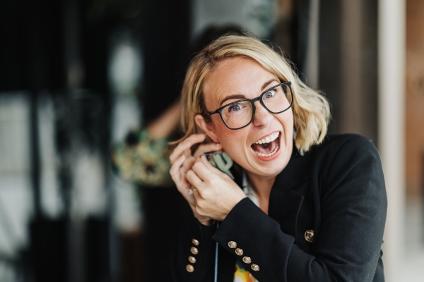 Woman laughing while putting on cactus-shaped earrings, wearing glasses and a blazer.