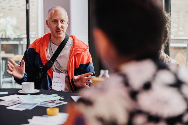 Leigh, wearing a bright red and navy jacket, speaks animatedly during a roundtable session at Design Leadership Melbourne 2024, seated with name tag visible.